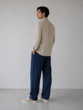 Man wearing a beige shirt and blue pants standing against a plain white wall.