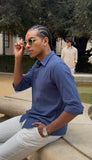 Man wearing a blue shirt and white pants sitting outdoors with sunglasses.