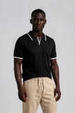 Man wearing a black polo shirt with white trim and beige pants on a light gray background