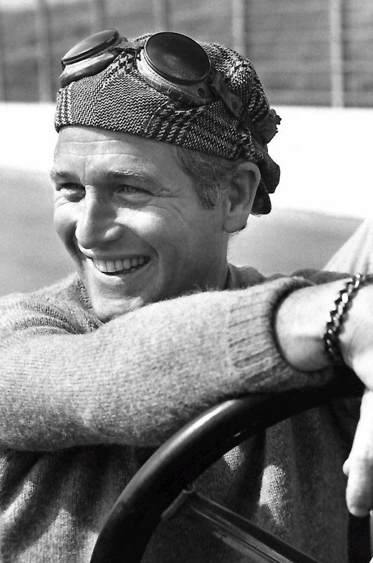 Paul Newman sitting at a racing car steering wheel, smiling, wearing a knit cap with goggles and a chunky sweater, 1971