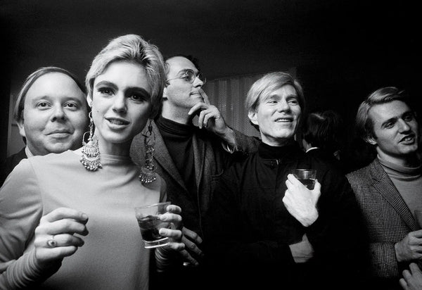 Andy Warhol wearing a black turtleneck at a social gathering, surrounded by a group of people, 1960s black and white photography