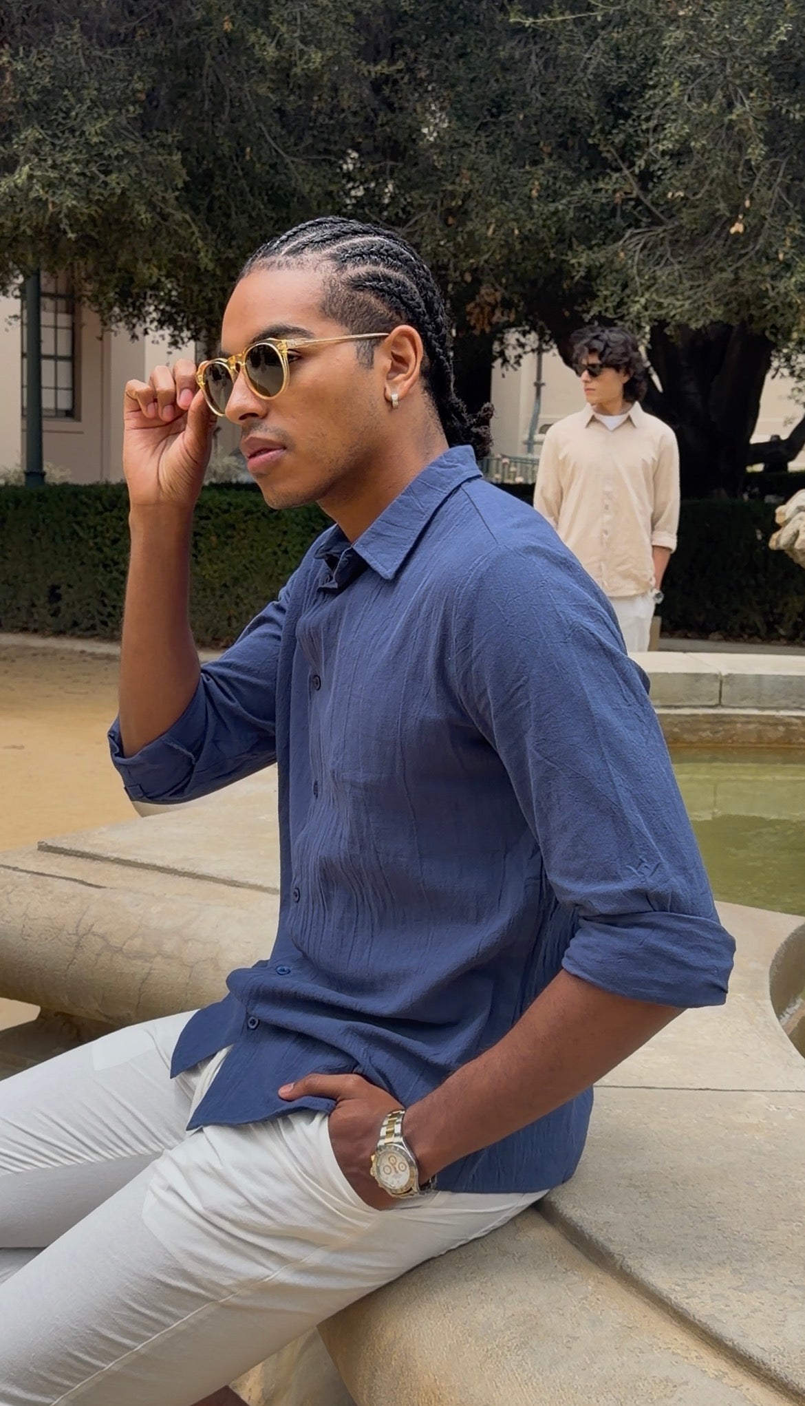 Man wearing a blue shirt and white pants sitting outdoors with sunglasses.
